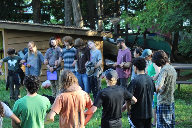 A group of people, appearing to be young adults, are standing in a circle outdoors. They seem to be engaged in some sort of group activity or discussion. The setting includes a wooden structure, possibly a shed or cabin, and trees in the background, suggesting a camp or retreat environment.